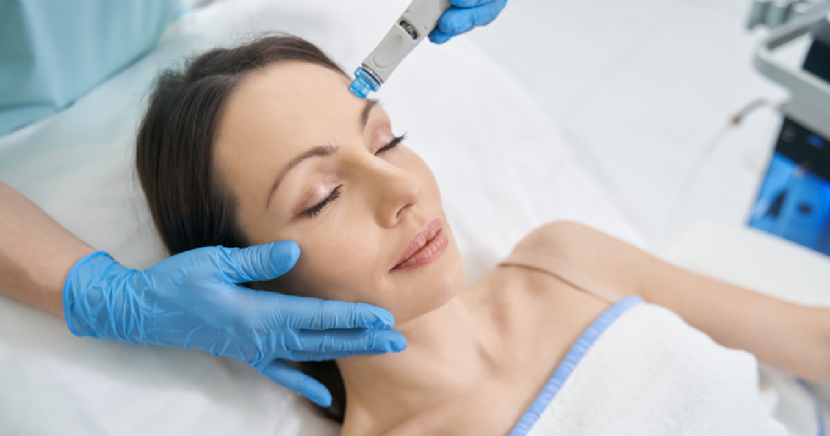 Woman receiving a HydraFacial treatment, lying peacefully with eyes closed, as a professional in blue gloves uses a handheld device on her forehead in Santa Rosa Beach & Destin