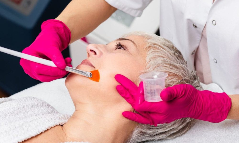 Beautiful senior woman having chemical peeling beauty treatment. The expert beautician is applying chemical solution onto woman's face.